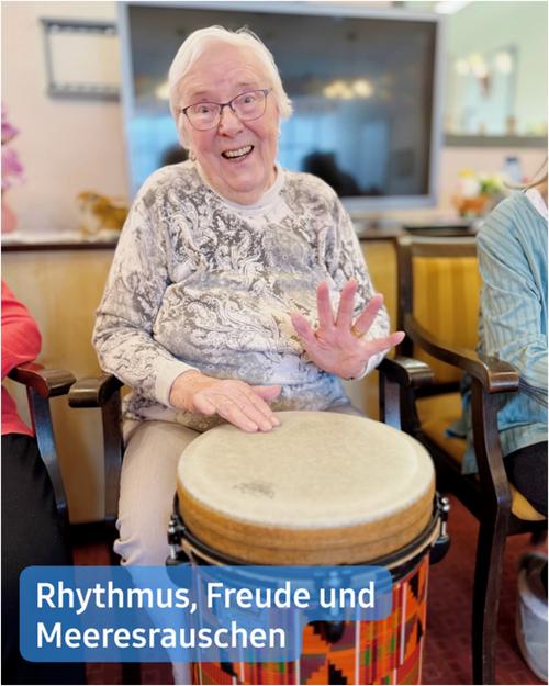 Nahaufnahme einer älteren Frau mit kurzen, weißen Haaren und Brille, die in einem Stuhl sitzt und ein Djembe-Trommel spielt. Sie trägt ein langärmliges, leicht graues, gemustertes Shirt. Ihr Gesichtsausdruck strahlt Freude aus, während eine Hand auf der Trommel ruht und die andere leicht erhoben ist, in einer einladenden Geste.  Der Hintergrund zeigt einen Gemeinschaftsraum oder einen Aufenthaltsbereich, möglicherweise in einer Seniorenresidenz oder einem Altenheim. Sichtbare Möbel umfassen Stühle und einen großen Flachbildfernseher. Im unteren rechten Bildbereich ist ein Teil einer anderen Person zu sehen, die ein türkisfarbenes Shirt trägt.  Am unteren Rand des Bildes befindet sich eine Bildunterschrift:  „Rhythmus, Freude und der Klang des Ozeans“