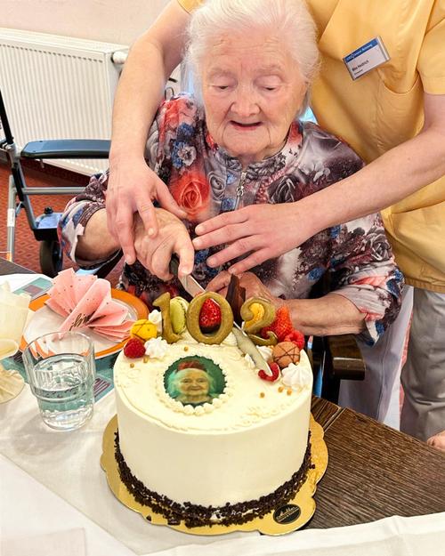 Eine Nahaufnahme zeigt eine Frau, möglicherweise in ihren 100ern, die lächelnd sitzt, während sie beim Schneiden einer mit der Zahl "102" in Gold dekorierten Torte unterstützt wird.  Der Sahnetorte ist weiß, mit Schokoladenraspeln um den Rand, und ziert ein essbares Bild sowie Früchte als Dekoration. Die Dame trägt ein blumiges Kleid und wird von einer Betreuerin assistiert, die hinter ihr positioniert ist und sanft ihre Hand zum Messer führt. Die Betreuerin trägt ein gelbes Oberteil. Ein kleinerer Teil der Umgebung ist ebenfalls sichtbar, darunter ein Tisch, ein Glas Wasser und Servietten.  Die gesamte Atmosphäre strahlt Feierlichkeit und liebevolle Fürsorge aus. Das Licht ist weich und warm und hüllt die Szenerie in einen angenehmen Glanz.